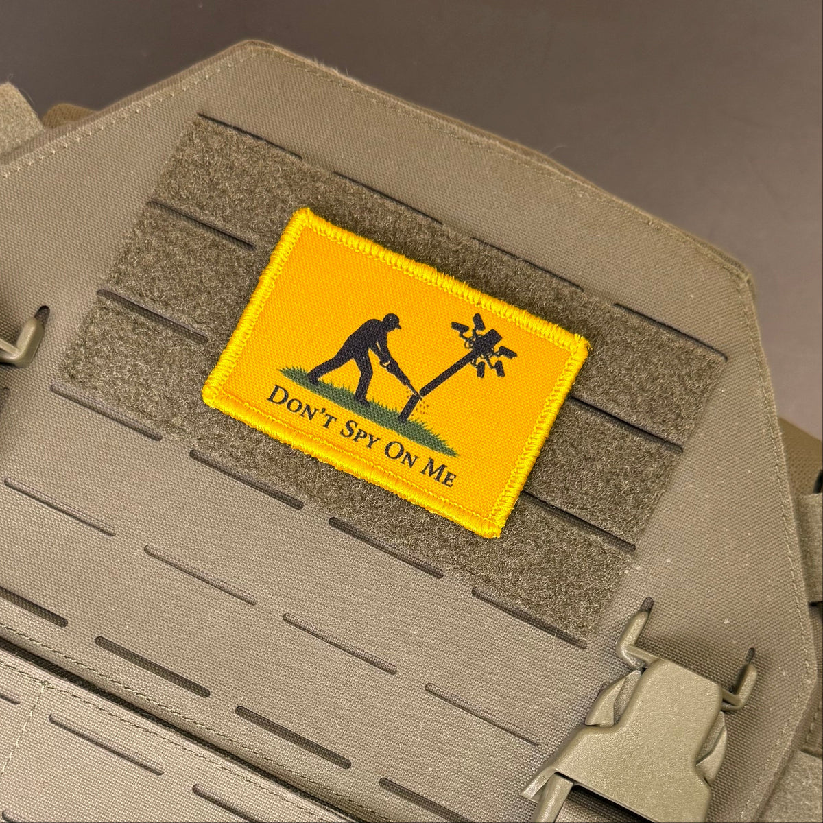 Don't Spy On Me printed canvas morale patch, a rectangular yellow tactical patch with a black silhouette of a silhouetted person in the artwork and the design letteringT SPY ON ME', attached to a tan webbing strap on a.