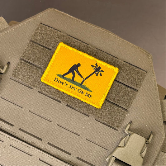 A rectangular yellow tactical patch with a black silhouette of a person planting a tree and the text 'DON'T SPY ON ME', attached to a tan webbing strap on a tactical vest.