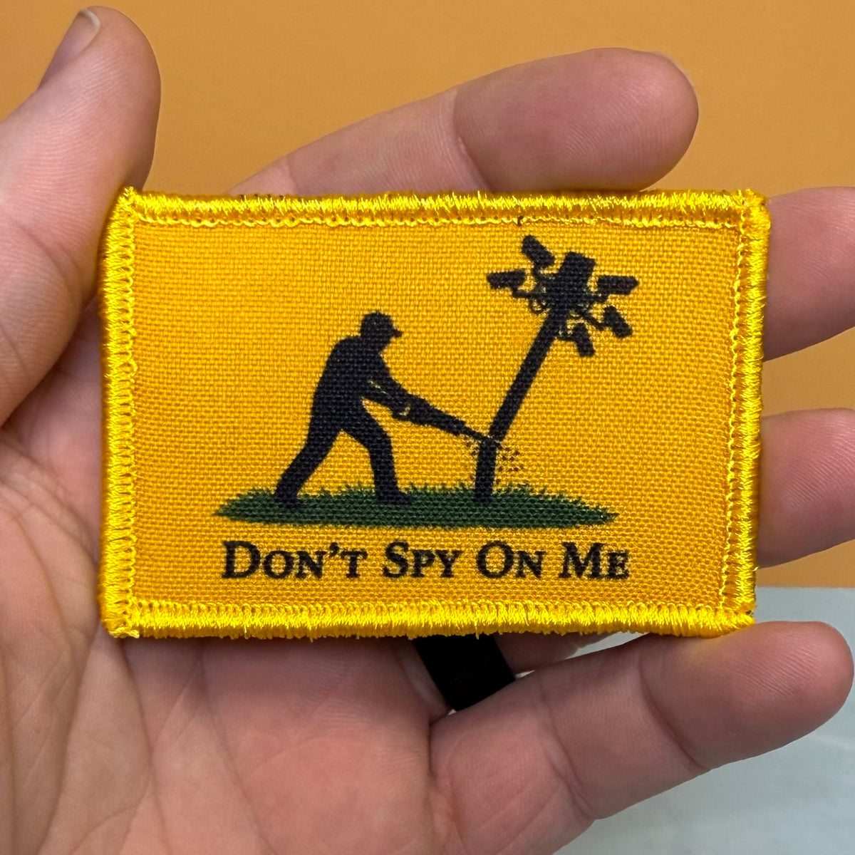 Don't Spy On Me printed canvas morale patch, a rectangular yellow printed patch held in a hand, featuring a black silhouette of a person cutting down a wire pole with the design letteringT SPY ON ME' below.