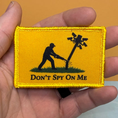 Don't Spy On Me printed canvas morale patch, a rectangular yellow printed patch held in a hand, featuring a black silhouette of a person cutting down a wire pole with the design letteringT SPY ON ME' below.