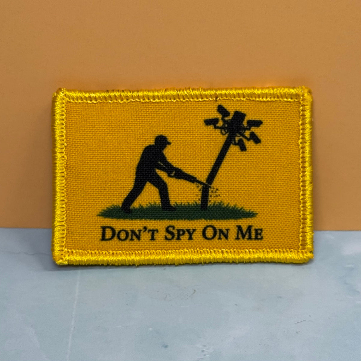 Don't Spy On Me printed canvas morale patch, silhouette of a person cutting down a wire pole, with the design letteringT SPY ON ME' below, shown against a tan and gray background.