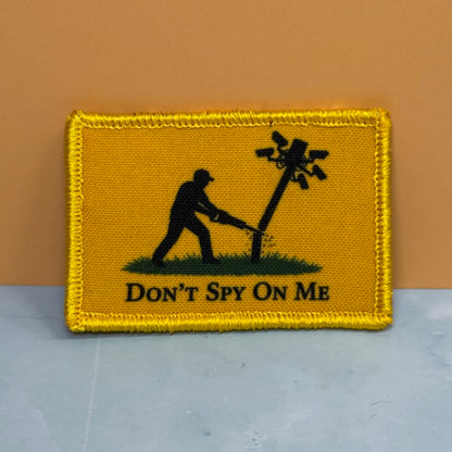 Don't Spy On Me printed canvas morale patch, silhouette of a person cutting down a wire pole, with the design letteringT SPY ON ME' below, shown against a tan and gray background.