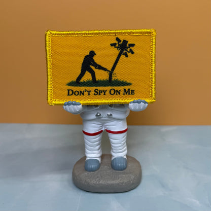 Don't Spy On Me printed canvas morale patch, raphic the design letteringT SPY ON ME' is held by a white astronaut figurine on a gray base against a warm tan and pale blue background.