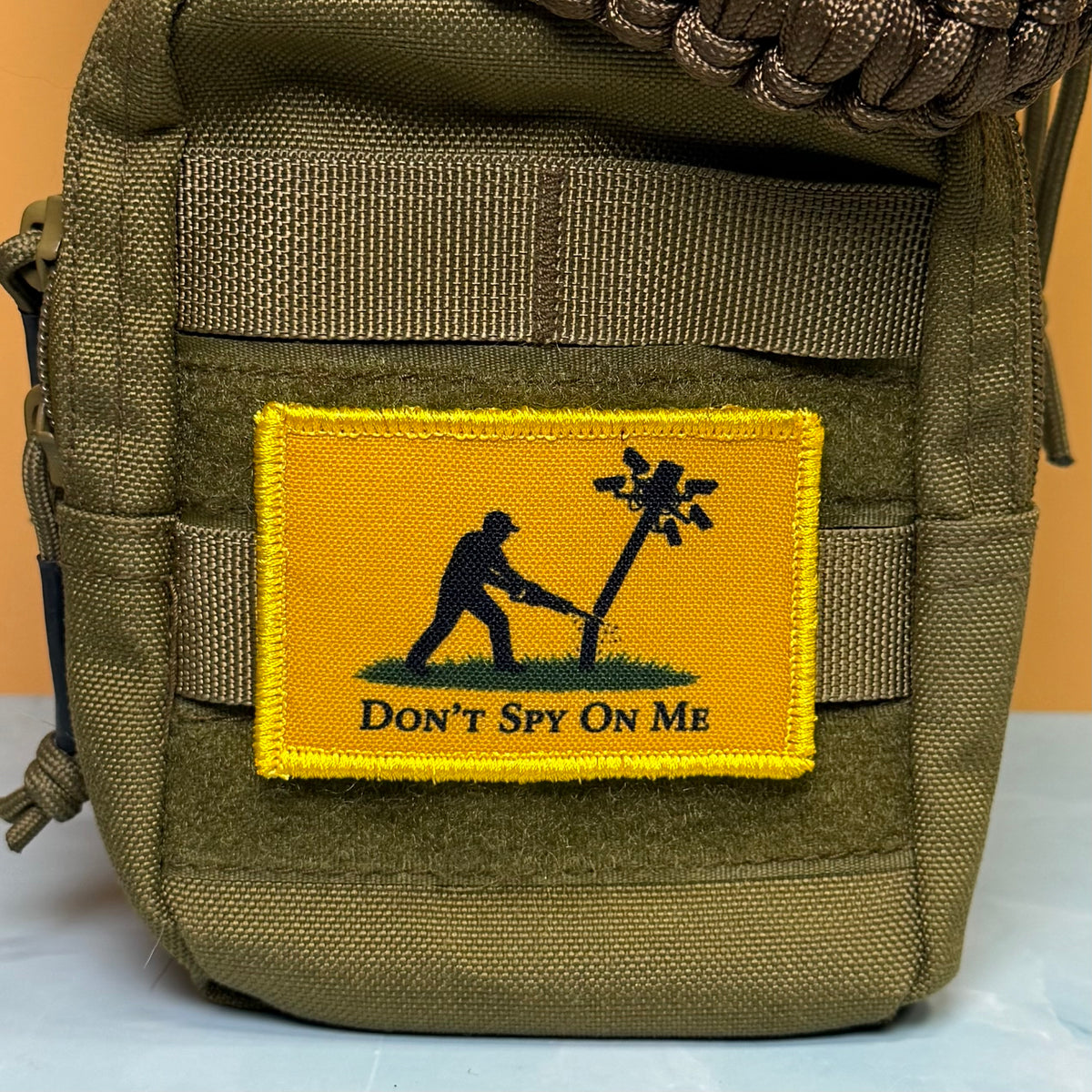 Don't Spy On Me printed canvas morale patch, an olive green tactical pouch featuring a rectangular yellow patch with a black silhouette of a person cutting down a pole, with the design letteringT SPY ON ME' below. A brown.