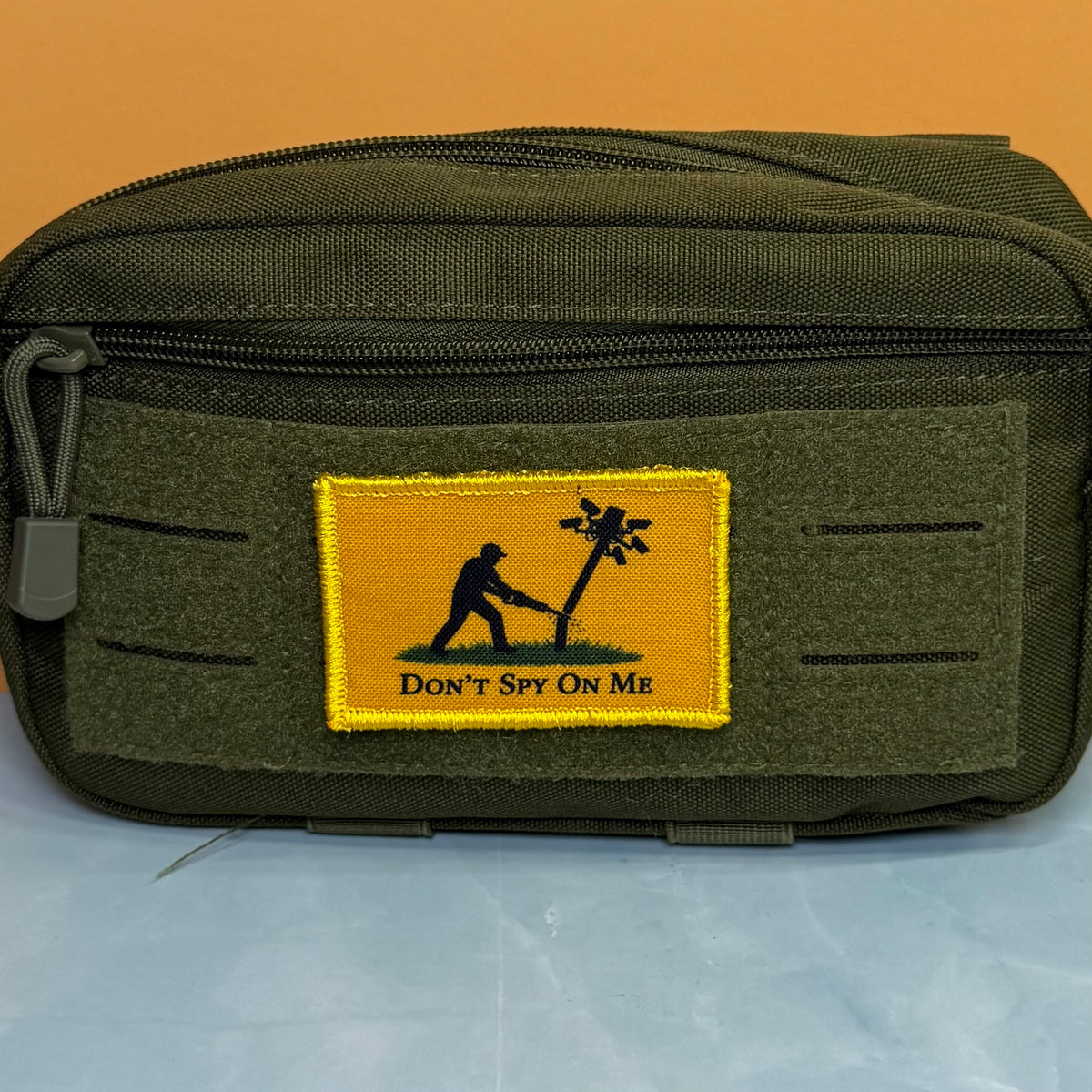 Don't Spy On Me printed canvas morale patch, an olive green tactical pouch with a rectangular yellow patch featuring a black silhouette of a person with a radio and the design letteringT SPY ON ME'.