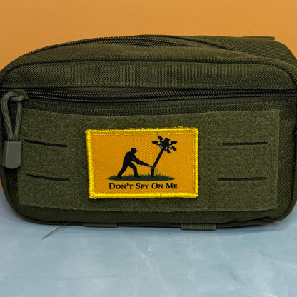 Don't Spy On Me printed canvas morale patch, an olive green tactical pouch with a rectangular yellow patch featuring a black silhouette of a person with a radio and the design letteringT SPY ON ME'.