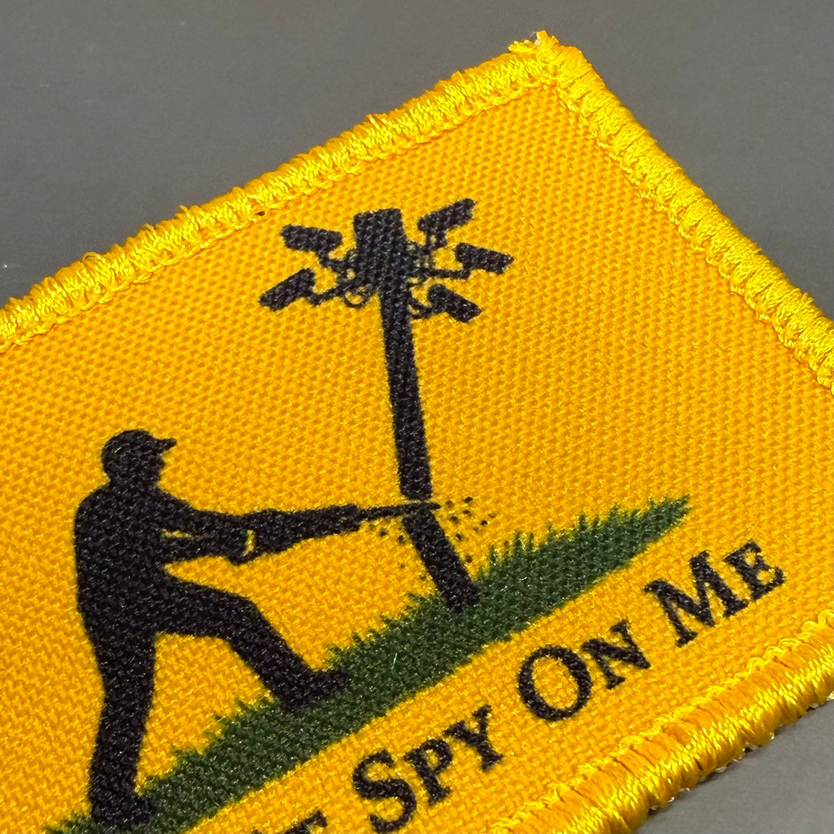 Don't Spy On Me printed canvas morale patch, a close-up of a yellow printed patch with a black silhouette of a person aiming a rifle at a utility pole, green grass, and black text the design lettering on a gray background.