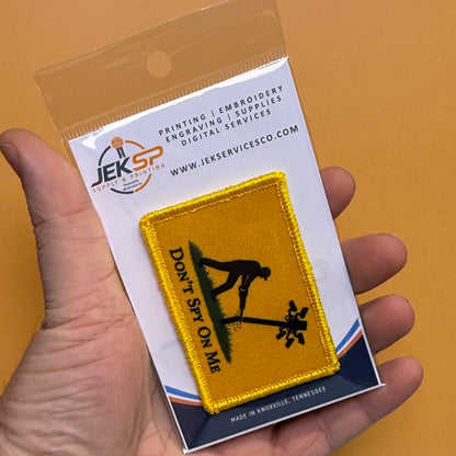 Don't Spy On Me printed canvas morale patch, a hand holds a packaged printed patch against a solid orange background. The patch is yellow with a black silhouette of a spy and the design letteringT SPY ON ME' on a green.
