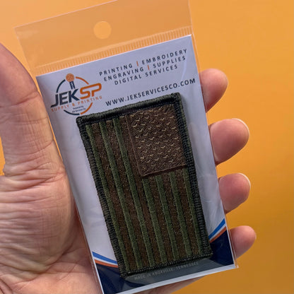 Subdued FDE Tactical American Flag embroidered morale patch: hand holding a packaged embroidered American flag patch. The patch is olive green and dark brown, and it is mounted on a white backing card with the JEKSP logo and text.
