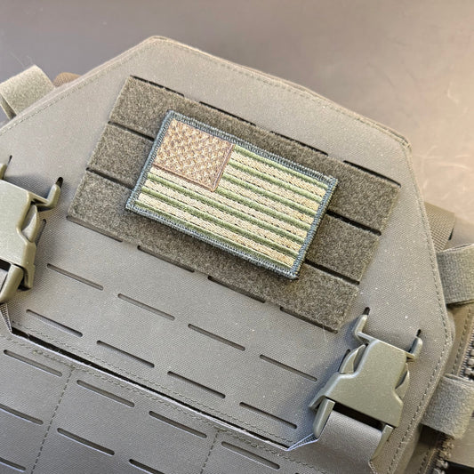 A close-up of a tactical patch featuring an American flag design in muted olive green, tan, and gray, mounted on a gray Velcro strip over a gray tactical vest or plate carrier.