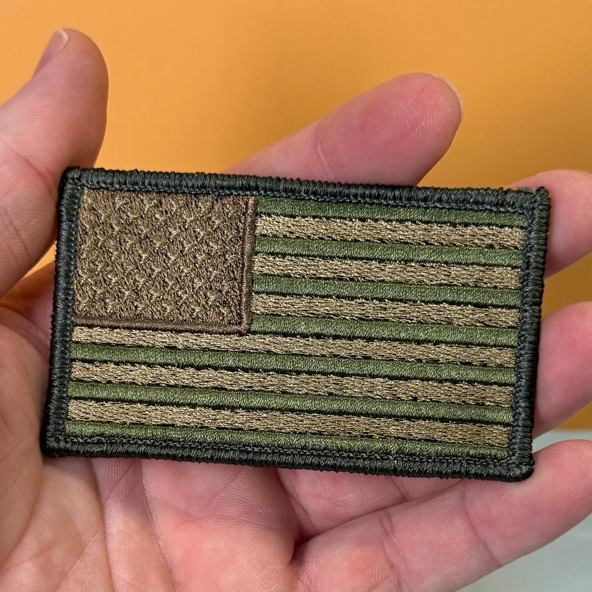 Subdued FDE Tactical American Flag embroidered morale patch: embroidered American flag patch in olive drab and tan, held in a person's hand against a solid orange background.