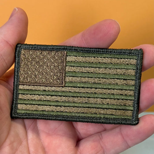 A rectangular embroidered American flag patch in olive drab and tan, held in a person's hand against a solid orange background.