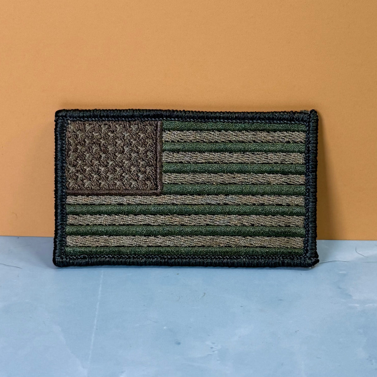 Subdued FDE Tactical American Flag embroidered morale patch: American flag patch in olive green, tan, and black, displayed on a light blue surface against a solid orange background.