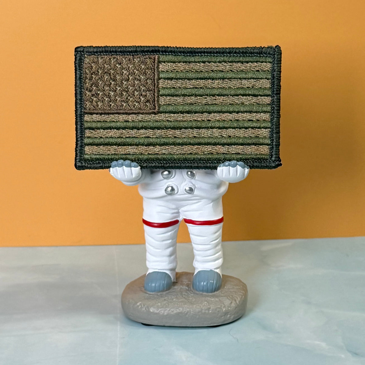 Subdued FDE Tactical American Flag embroidered morale patch: small astronaut figurine holding a rectangular American flag patch with a camouflage pattern. The astronaut is white with red stripes on the legs and gray boots, standing on a gray.