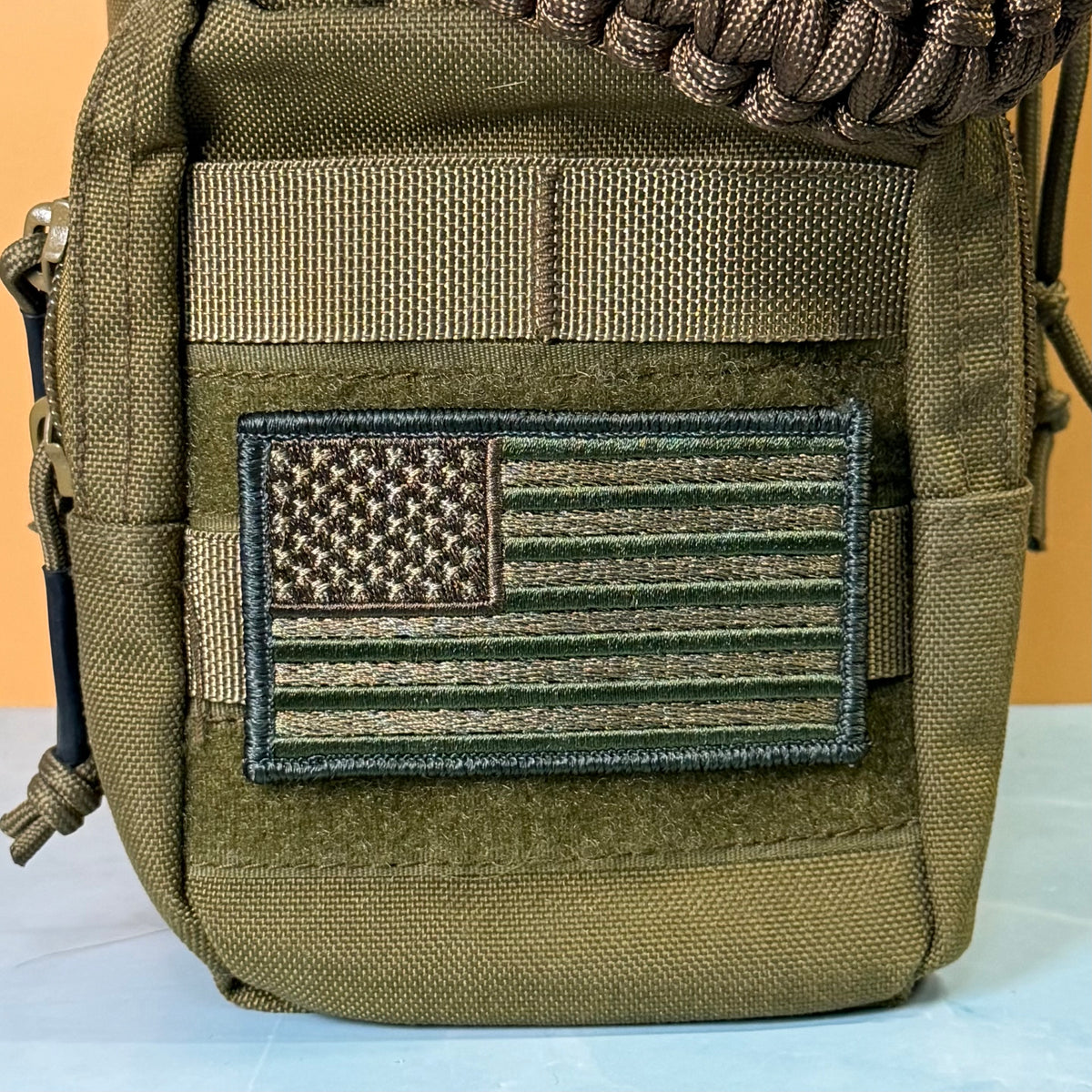 Subdued FDE Tactical American Flag embroidered morale patch: olive drab tactical pouch featuring a rectangular embroidered American flag patch in matching olive and brown tones, with a tan webbing strap and a brown paracord detail at the top.