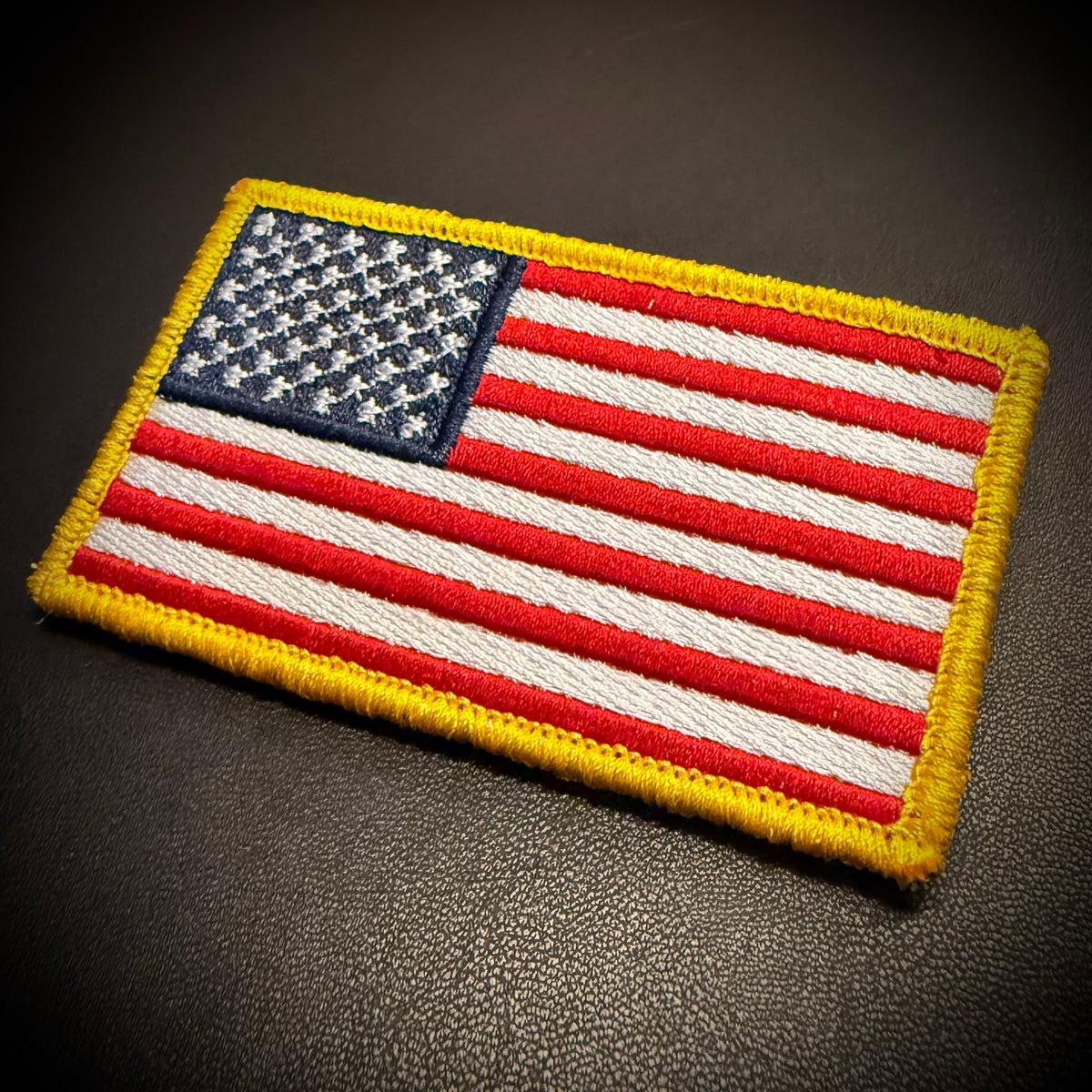 An embroidered American flag patch with red and white stripes, a blue field with white stars, and a yellow border, shown on a dark textured surface.