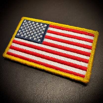 An embroidered American flag patch with red and white stripes, a blue field with white stars, and a yellow border, shown on a dark textured surface.