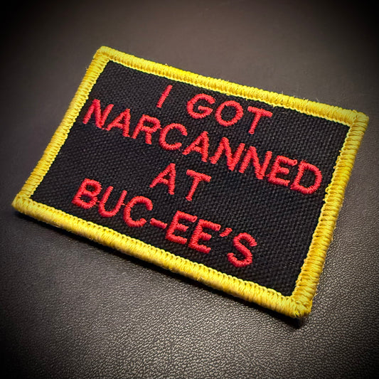 A rectangular embroidered patch with a black background, yellow border, and red text reading 'I GOT NARCANED AT BUC-EE'S', shown on a dark textured surface.