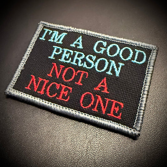 A rectangular embroidered patch with a black background and gray stitched border, featuring the text 'I'M A GOOD PERSON NOT A NICE ONE' in light blue and red lettering, placed on a dark textured surface.