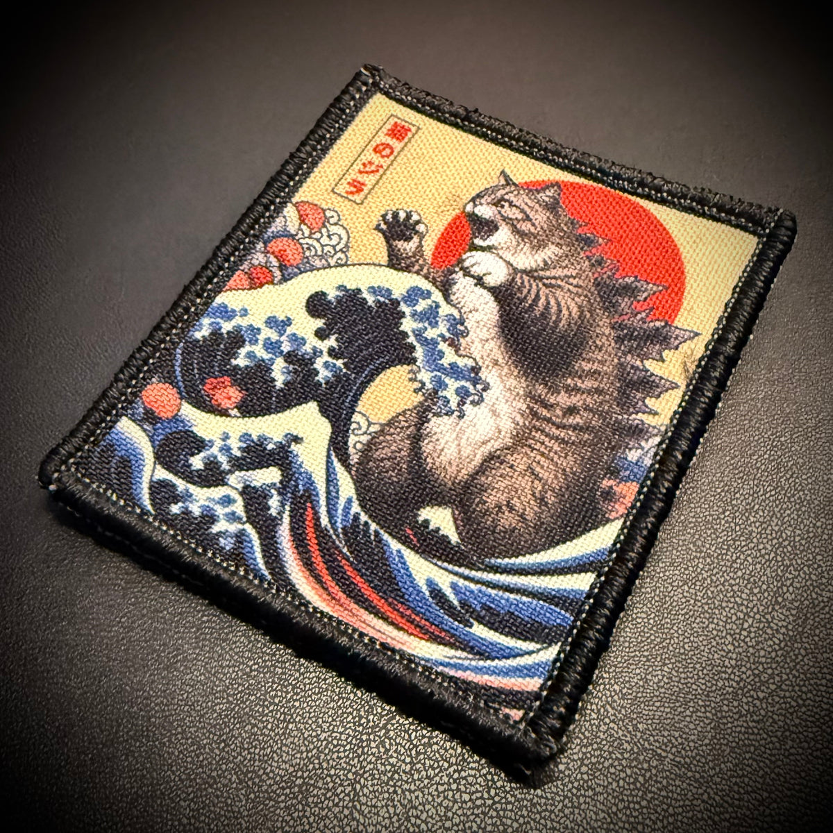 A square embroidered patch featuring a tabby cat in a Japanese wave-and-sun style print, photographed on a dark textured surface.