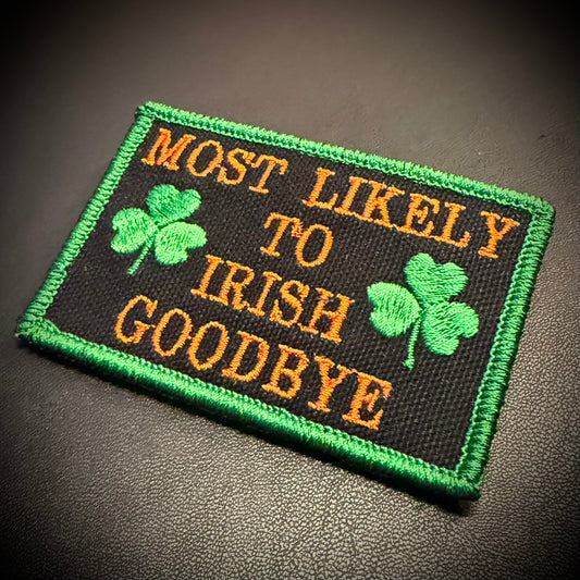 A rectangular embroidered patch with a black background, green border, orange text reading 'MOST LIKELY TO IRISH GOODBYE,' and two green shamrocks, photographed on a dark textured surface.