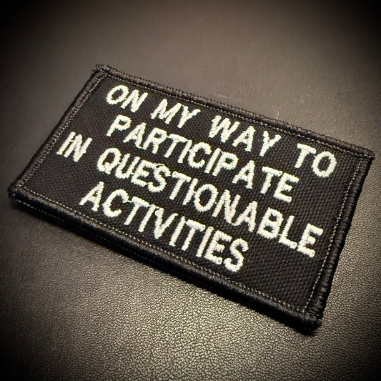 A rectangular black embroidered patch with white text reading 'ON MY WAY TO PARTICIPATE IN QUESTIONABLE ACTIVITIES' on a dark textured surface.