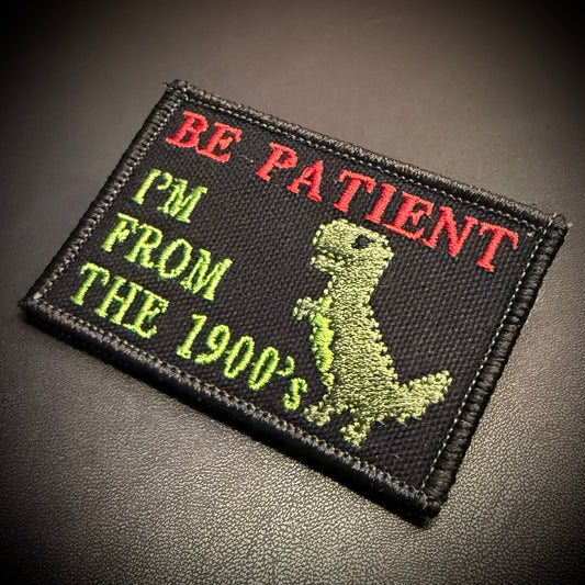 A rectangular embroidered patch on a dark textured background. The patch is black with red and green embroidered text reading 'BE PATIENT I'M FROM THE 1900's' and a green dinosaur graphic.