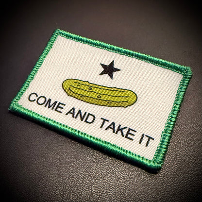A rectangular embroidered patch with a green border, featuring a yellow pickle, a black star, and the text 'COME AND TAKE IT' on a white background, photographed on a dark textured surface.