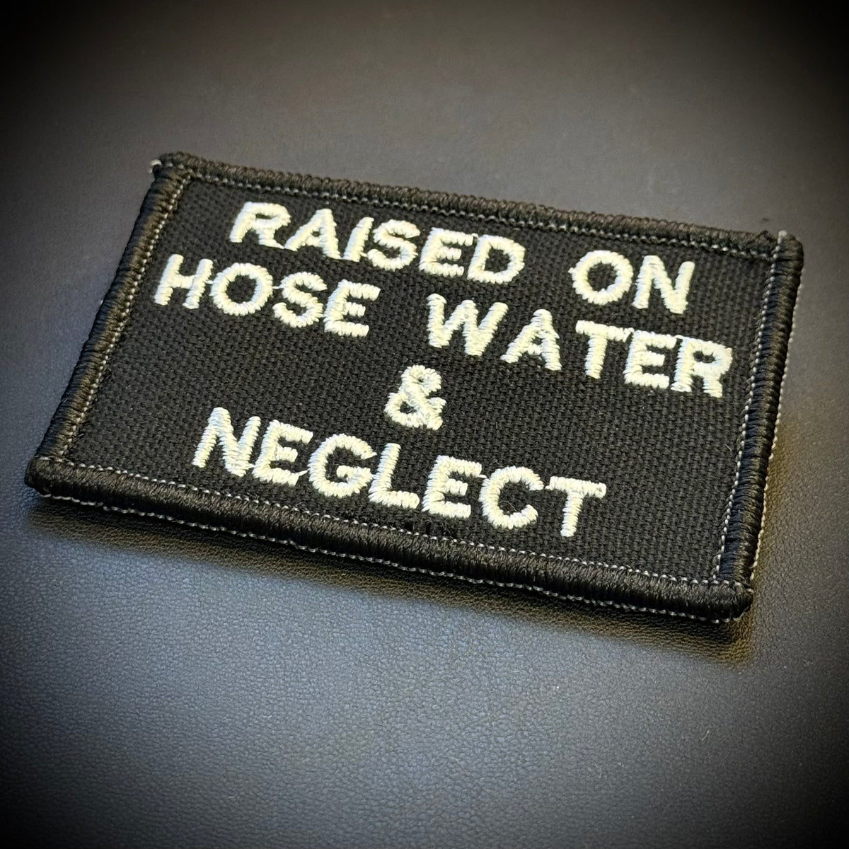 A rectangular black embroidered patch with white text reading 'RAISED ON HOSE WATER & NEGLECT' on a dark gray background.