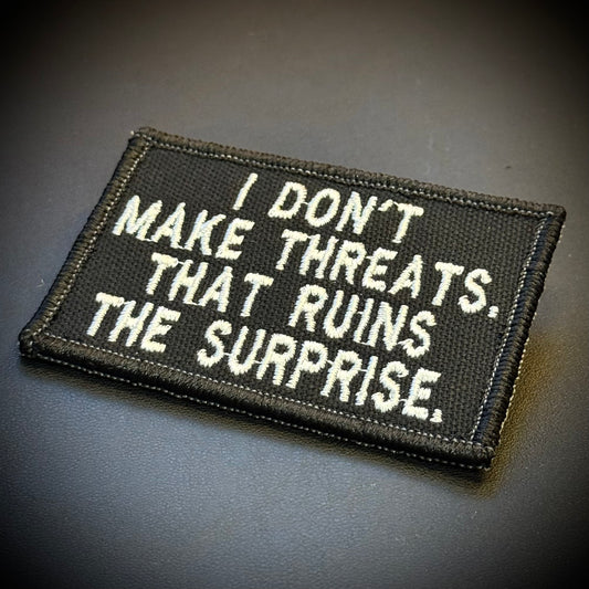 I Don't Make Threats, That Ruins The Surprise embroidered morale patch: black embroidered patch with white text reading 'I DON'T MAKE THREATS. THAT RUINS THE SURPRISE.