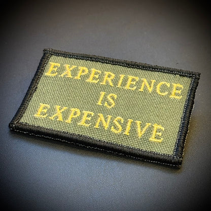 An embroidered rectangular patch with the phrase 'EXPERIENCE IS EXPENSIVE' in yellow lettering on an olive green background, finished with a black stitched border, shown on a dark gray surface.