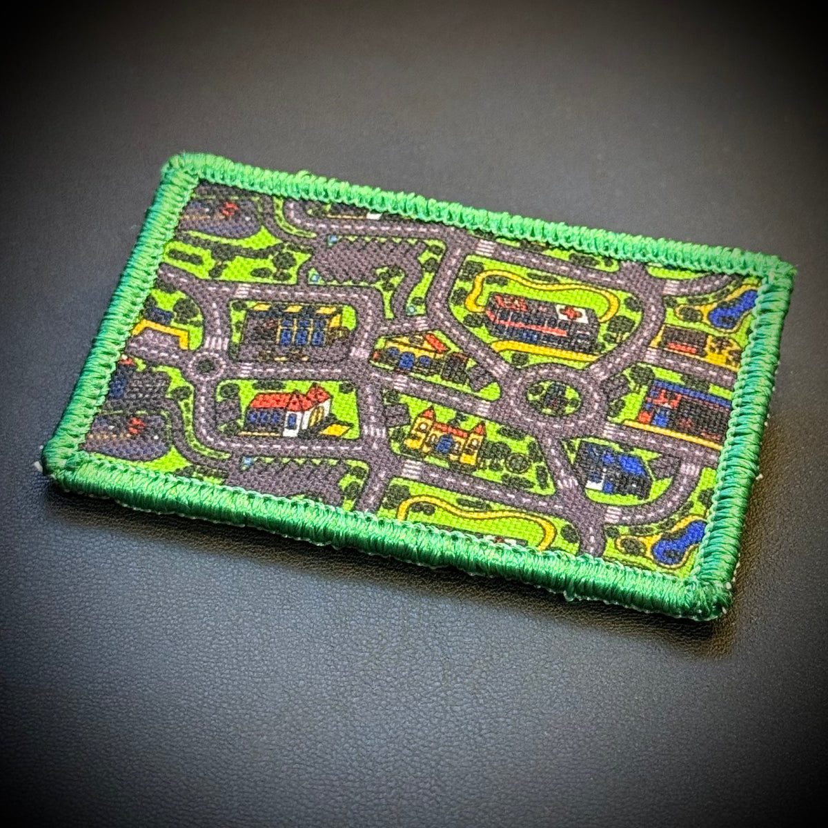 A rectangular embroidered patch featuring a colorful street map design with a bright green stitched border, placed on a dark textured background.