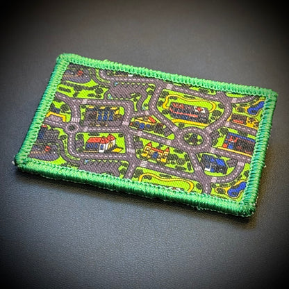 A rectangular embroidered patch featuring a colorful street map design with a bright green stitched border, placed on a dark textured background.