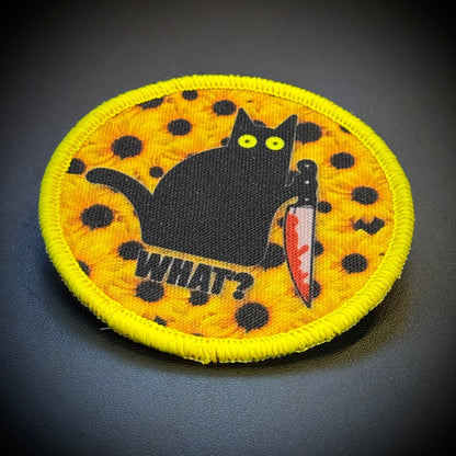 A circular embroidered patch featuring a black cat holding a knife, with the word 'WHAT?' below it, on a yellow background with black polka dots and a bright yellow stitched border.