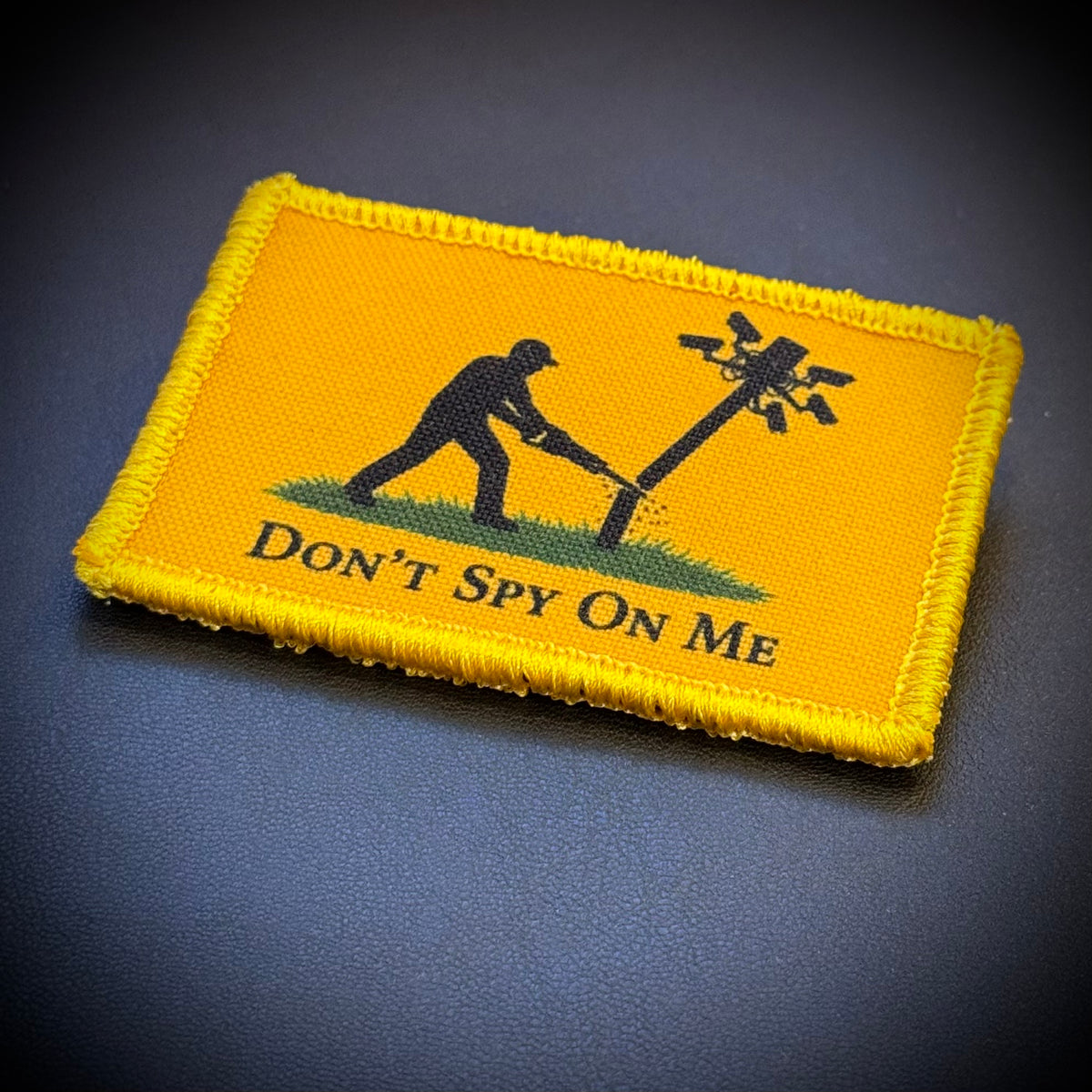 A rectangular embroidered tactical patch with a bright yellow border and yellow background, featuring a black silhouette of a person cutting down a wire pole with the text 'DON'T SPY ON ME' below. The patch is photographed on a dark textured surface.