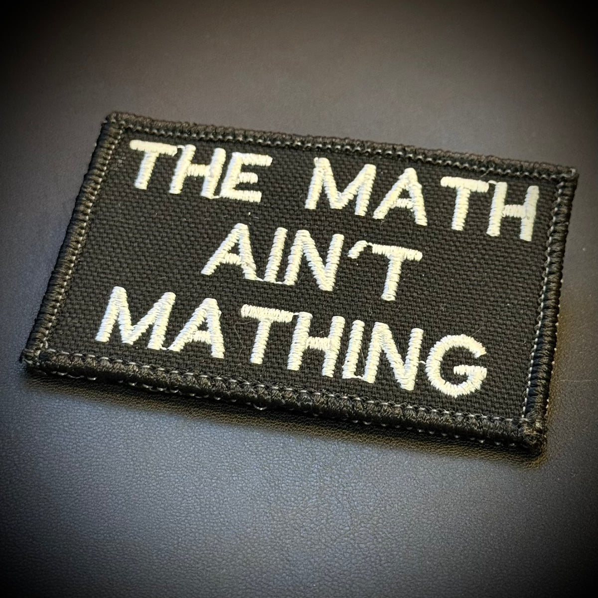 A rectangular black embroidered patch with white text reading 'THE MATH AIN'T MATHING' on a dark gray background.