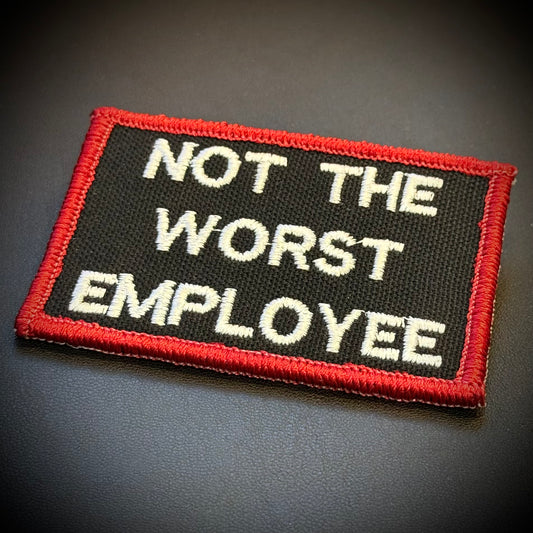A rectangular embroidered patch with a black background, red stitched border, and white text reading 'NOT THE WORST EMPLOYEE' on a dark surface.