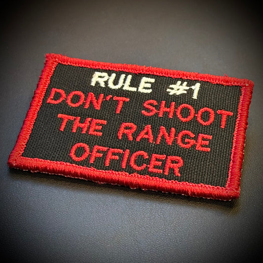 A rectangular embroidered patch with a black background, red border, and red and white text reading 'RULE #1 DON'T SHOOT THE RANGE OFFICER' on a dark surface.