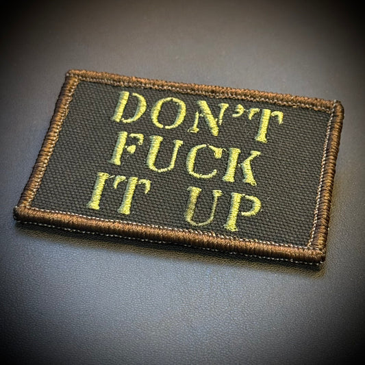 Don't Fuck It Up embroidered morale patch: with the text 'DON'T FUCK IT UP' in olive green on a black background, finished with a brown stitched border, leaning against a dark background.