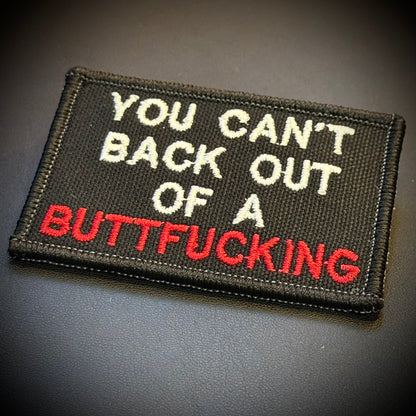 A rectangular embroidered patch with a black background and white and red text reading 'YOU CAN'T BACK OUT OF A BUTTFUCKING', shown on a dark gray surface.