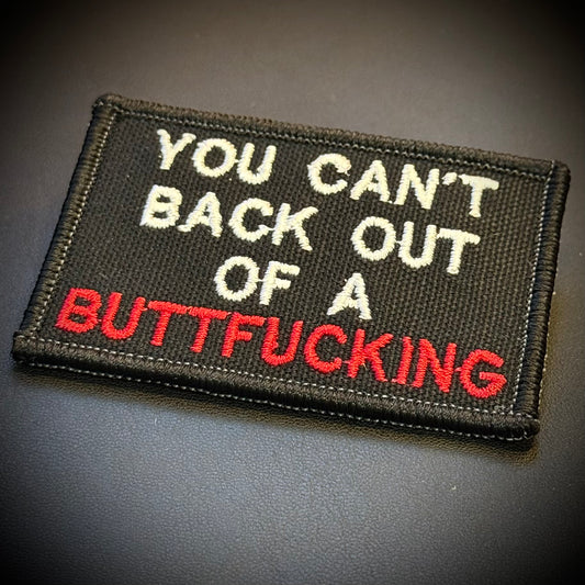 A rectangular embroidered patch with a black background and white and red text reading 'YOU CAN'T BACK OUT OF A BUTTFUCKING', shown on a dark gray surface.