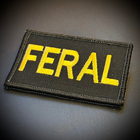 A rectangular black embroidered patch with the word 'FERAL' in bold yellow letters, shown on a dark gray textured background.