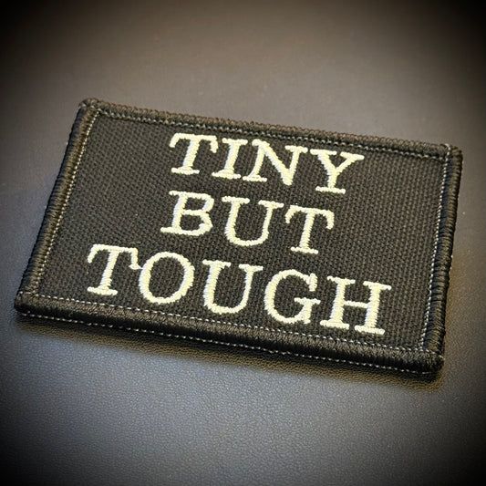 A rectangular embroidered patch with the words 'TINY BUT TOUGH' in cream lettering on a black fabric background, shown on a dark textured surface.