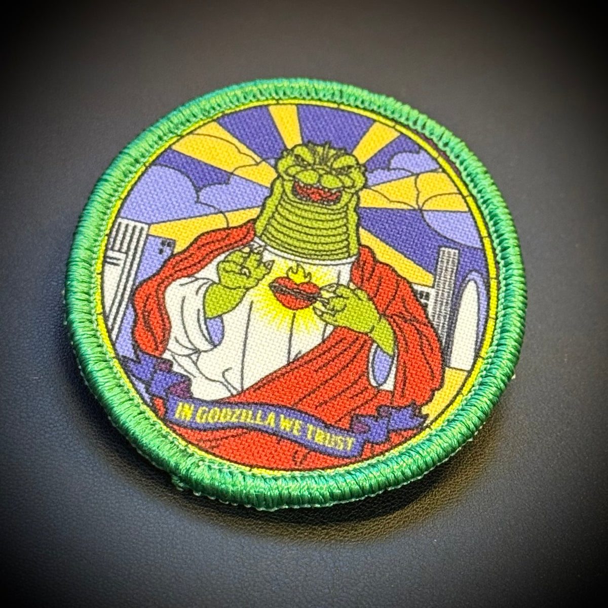 A circular embroidered patch featuring a green-skinned character in a red robe with the text 'IN GODZILLA WE TRUST' on a dark textured background.