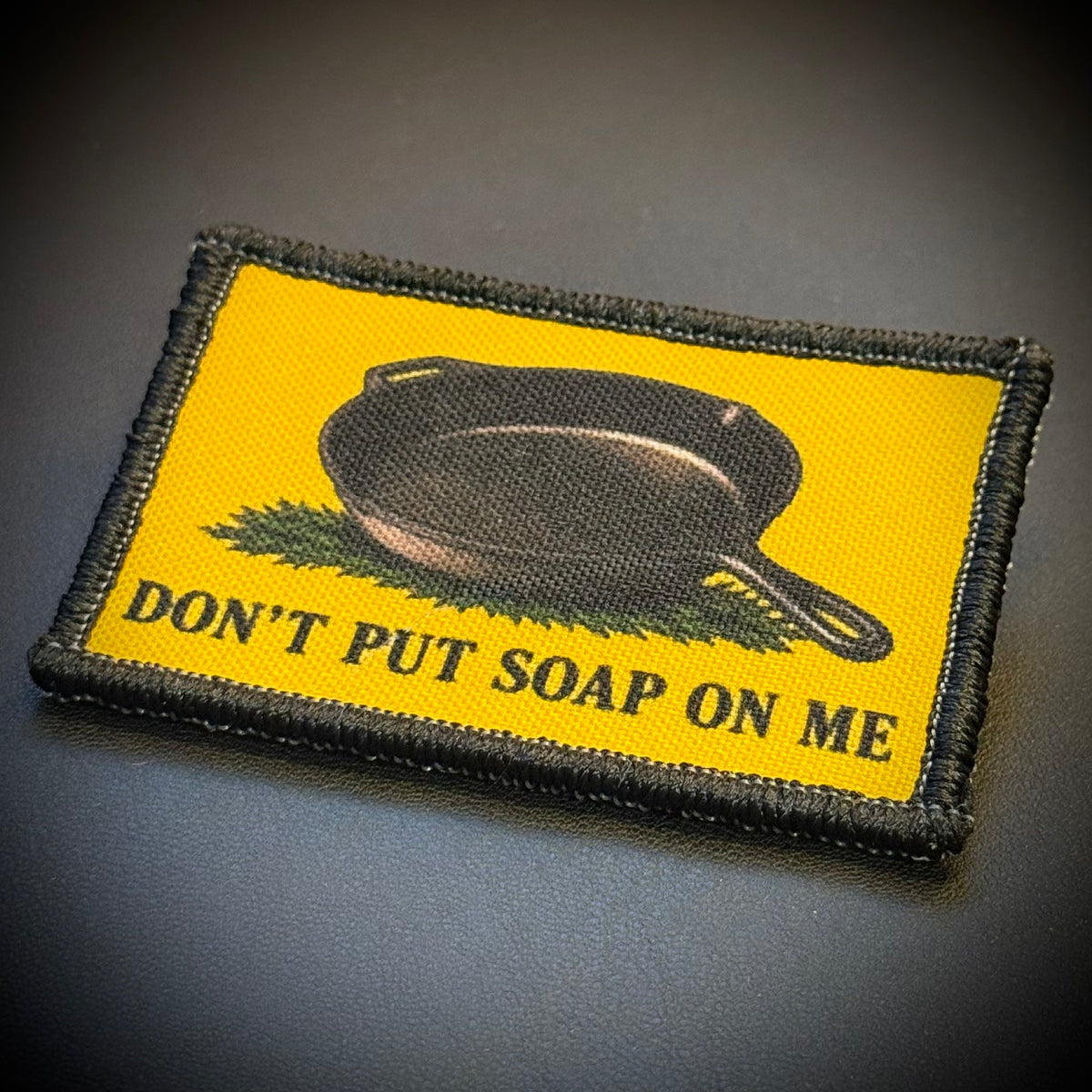 A rectangular embroidered patch with a yellow background, a black cast-iron skillet, green grass, and the text 'DON'T PUT SOAP ON ME' in black, shown on a dark gray surface.