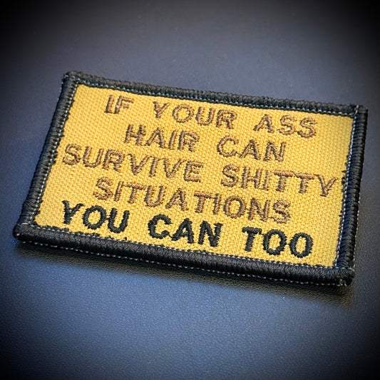 A rectangular embroidered morale patch with a yellow background and black border, featuring the humorous text 'IF YOUR ASS HAIR CAN SURVIVE SHITTY SITUATIONS YOU CAN TOO' in black and brown thread, shown on a dark textured background.