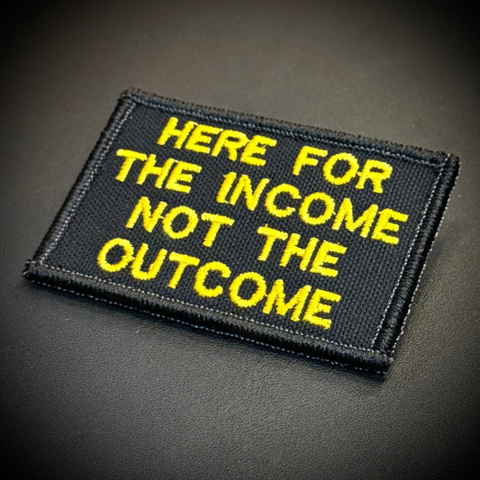 A rectangular black embroidered patch with yellow text reading 'HERE FOR THE INCOME NOT THE OUTCOME' on a dark textured background.