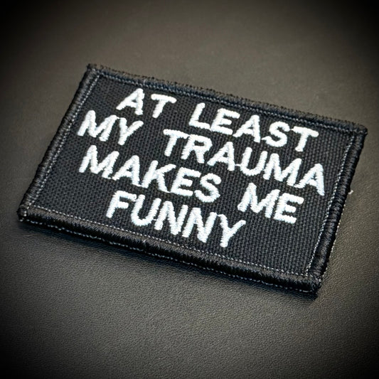 A rectangular black embroidered patch with white text reading 'AT LEAST MY TRAUMA MAKES ME FUNNY' on a dark textured background.
