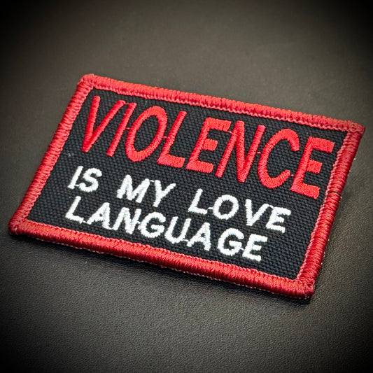 An embroidered rectangular patch with a black background, red border, and red and white text reading 'VIOLENCE IS MY LOVE LANGUAGE', photographed on a dark textured surface.