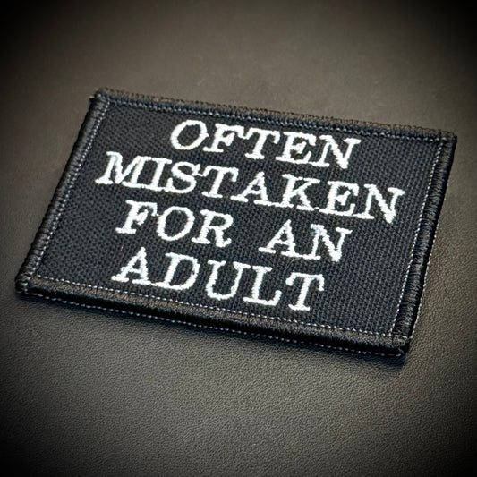 A rectangular black embroidered patch with white text reading 'OFTEN MISTAKEN FOR AN ADULT' on a dark textured background.